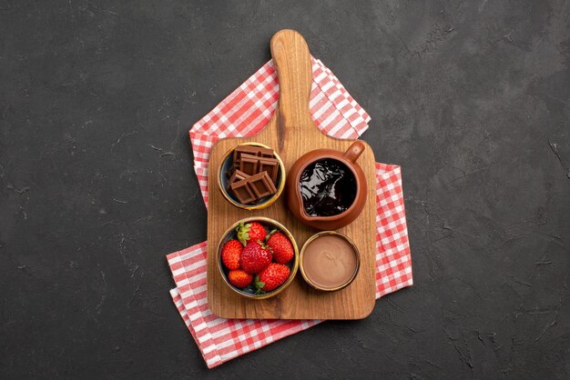 Draufsicht aus der Ferne Dessert auf Tischdeckenschalen mit appetitlicher Schokoladencreme und Erdbeeren auf dem Schneidebrett auf der rosa-weiß karierten Tischdecke auf dem dunklen Tisch