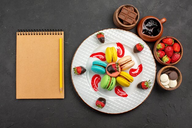 Draufsicht aus der Ferne Dessert leckere Makronen und Erdbeeren neben dem Sahnenotizbuch mit Bleistift und Schalen mit Schokoladenerdbeeren und Schokoladencreme auf dem Tisch