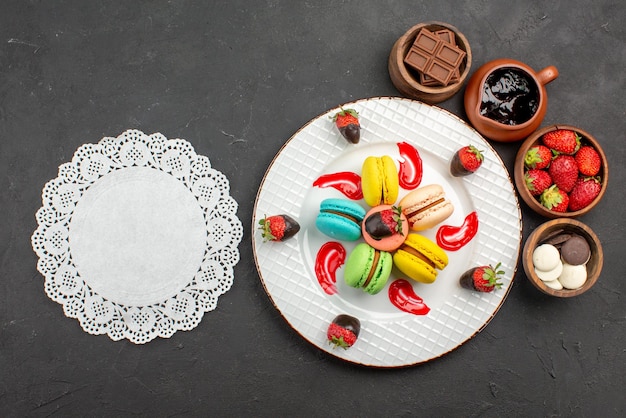 Draufsicht aus der Ferne Dessertmakronen und Erdbeeren im Teller neben dem Spitzendeckchen und Schalen mit Schokoerdbeeren und Schokocreme auf dem Tisch