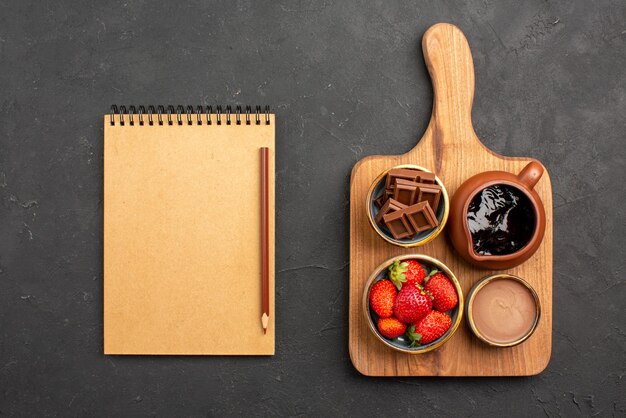 Draufsicht aus der Ferne Dessertschalen mit appetitlicher Schokoladencreme und Erdbeeren auf dem Schneidebrett neben dem Sahnenotizbuch und Bleistift auf dem dunklen Tisch