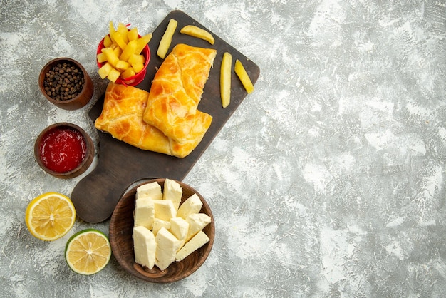 Draufsicht aus der Ferne Fastfood zwei Kuchen und Pommes Frites auf dem Brett neben den Schüsseln mit Käseketchup und schwarzer Pfefferzitrone auf dem grauen Tisch