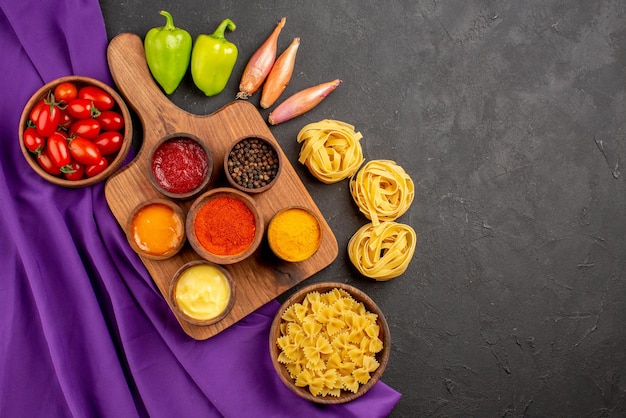 Draufsicht aus der ferne gewürze und nudeln drei arten von gewürzen und saucen auf dem holzbrett neben der schüssel mit nudeln grüne kugelpfefferzwiebel und tomaten auf der lila tischdecke auf dem dunklen tisch