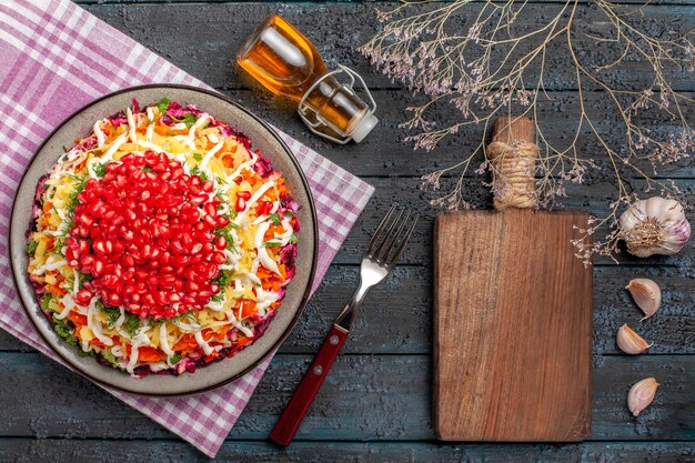 Draufsicht aus der Ferne Granatapfel in der Tellerschale auf der karierten Tischdecke neben dem Schneidebrett Gabel Äste Knoblauchflasche Öl auf dem Tisch