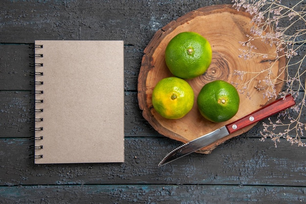 Kostenloses Foto draufsicht aus der ferne grüne limetten auf braunem holzbrett neben messernotizbuch und ästen auf dem grauen tisch