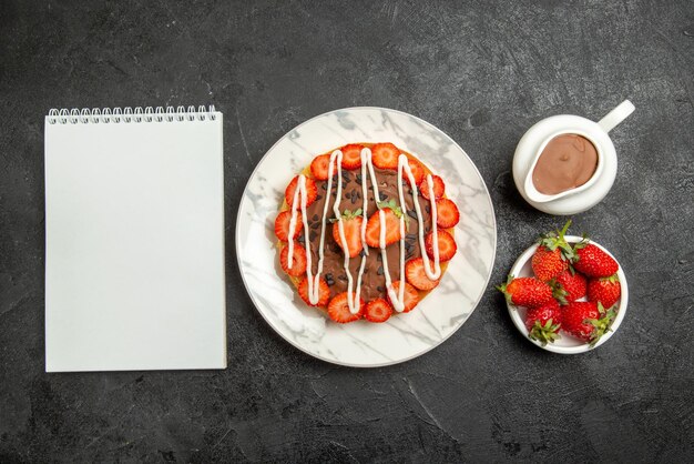 Draufsicht aus der Ferne Kuchen mit Beeren weiße Notizbuchschüsseln mit Erdbeeren und Schokoladencremetorte mit Erdbeeren und Schokolade auf dem Tisch