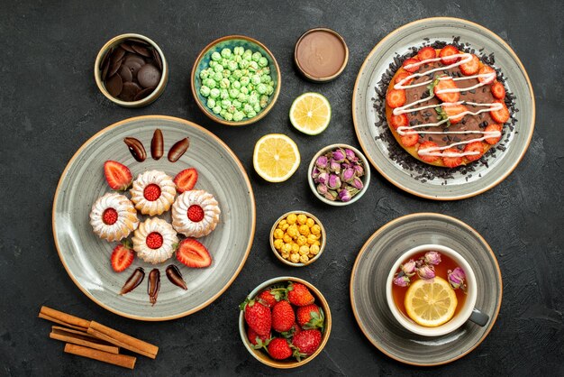 Draufsicht aus der Ferne Kuchen mit Süßigkeiten Kuchen mit Schokolade und Erdbeer Schwarztee Zitronen Teller mit Keksen mit Erdbeerschalen Schokolade und verschiedenen Süßigkeiten auf schwarzem Tisch