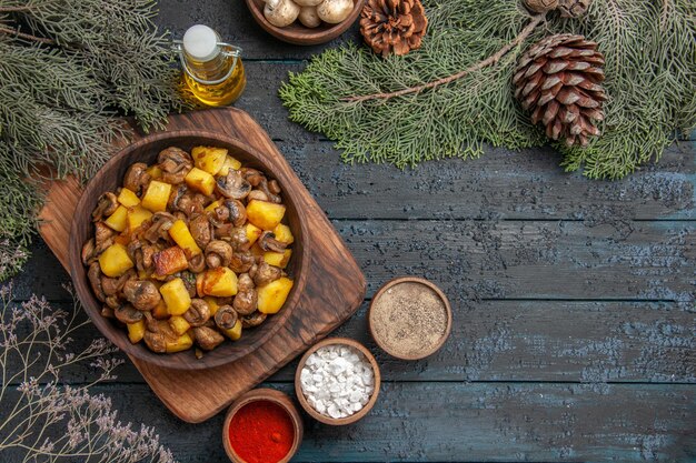 Draufsicht aus der Ferne Schüssel mit Speiseschüssel Kartoffeln mit Pilzen auf dem Schneidebrett neben den bunten Gewürzen unter Ölschüssel mit weißen Champignons und Fichtenzweigen