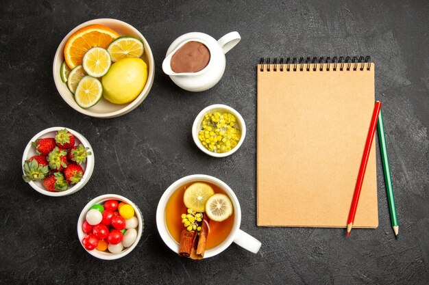 Draufsicht aus der Ferne Süßigkeiten auf dem Tisch eine Tasse Tee mit Zitronen- und Zimtstangen neben dem cremefarbenen Notizbuch mit grünem und rotem Bleistift und Schalen mit Beeren und Kräutern