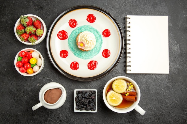 Draufsicht aus der Ferne Süßigkeiten mit Tee weißer Notebook Cupcake mit Sahne eine Tasse Kräutertee mit Zitrone neben den Schalen mit Schokoladencreme Erdbeeren und Bonbons