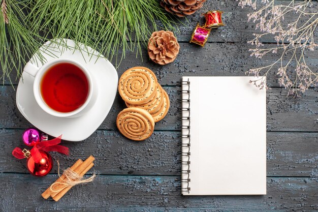 Draufsicht aus der Ferne Teefichte mit Zapfen und Weihnachtsspielzeug neben den Zimtstangen eine Tasse Tee auf der Untertasse und weißem Notizbuch