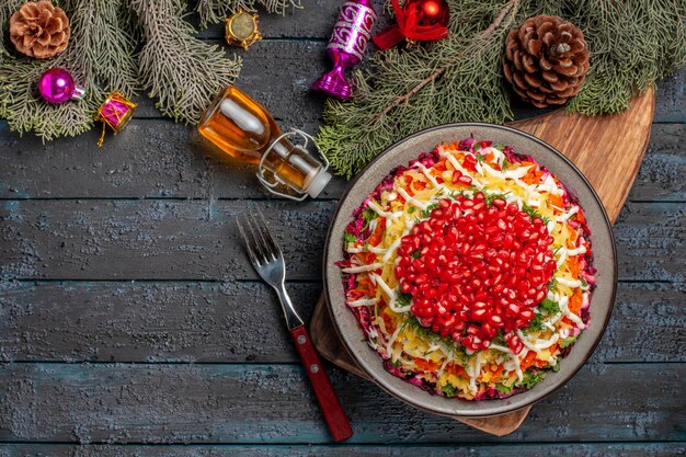 Draufsicht aus der Ferne Teller in der Tellerschale mit Granatapfel-Karotten auf dem Schneidebrett neben der Flasche Ölgabel und Ästen mit Zapfen und Weihnachtsbaumspielzeug