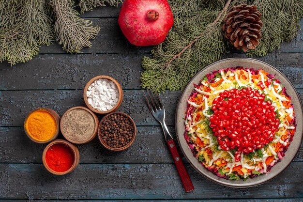 Draufsicht aus der Ferne Teller und Äste Schüsseln mit Gewürzgabel Granatapfel und Äste mit Zapfen neben dem Teller mit appetitlichen Granatapfel-Karotten und Kartoffeln