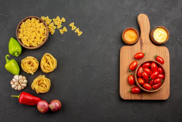 Draufsicht aus der Ferne Tomaten und Gewürze verschiedene Nudelsorten Paprika Zwiebel Knoblauch auf der linken Seite und die Schüssel Tomaten auf dem Holzbrett und Soßen auf der rechten Seite des Tisches