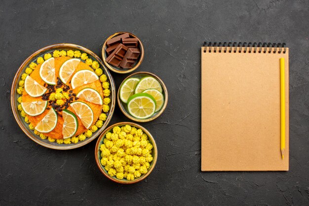 Draufsicht aus der Ferne Zitrusfrüchte und Schokoladen-Orangenkuchen mit Schokolade neben den Schalen mit Bonbonschokolade und Limettenscheiben neben dem Notizbuch und Bleistift auf dem schwarzen Tisch