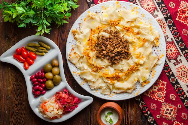 Draufsicht azerbaijani Guru khingal kaukasische Nudeln mit gebratenem gehacktem Fleisch und Zwiebeln mit Sauerrahmsauce und Gurken auf einer Tischdecke horizontal