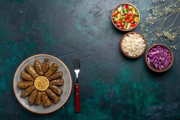 Draufsicht Blatt Dolma östliches Fleischmehl gerollt in grünen Blättern mit geschnittenem Gemüse auf blauem Schreibtischfleisch Abendessen Ostgericht