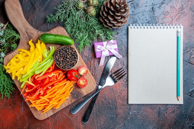 Draufsicht bunt geschnittene Paprika schwarzer Pfeffer Tomaten Gurke auf Schneidebrett Bleistift auf Notizblock Gabel und Messer kleines Geschenk auf dunkelrotem Tisch