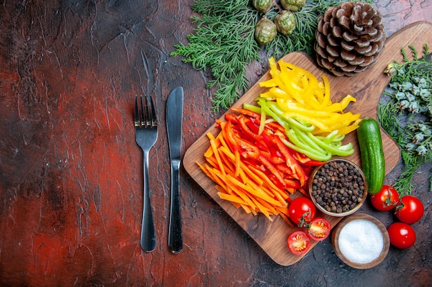 Draufsicht bunte geschnittene Paprika schwarzer Pfeffer Tomaten Gurke auf Schneidebrett Gabel und Messer Salz Kiefer Zweige auf dunkelroten Tisch freien Raum