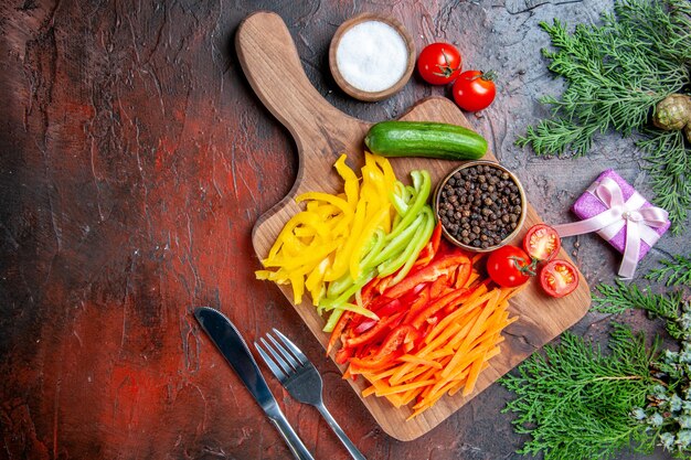 Draufsicht bunte geschnittene Paprika schwarzer Pfeffer Tomaten Gurke auf Schneidebrett Salzgabel und Messer kleines Geschenk auf dunkelrotem Tisch