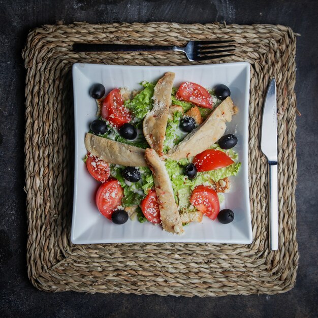 Draufsicht Caesar Salat mit Huhn, Tomate, Salat, Oliven, Cracker, Parmesankäse auf einem Weidenständer in einem weißen Teller