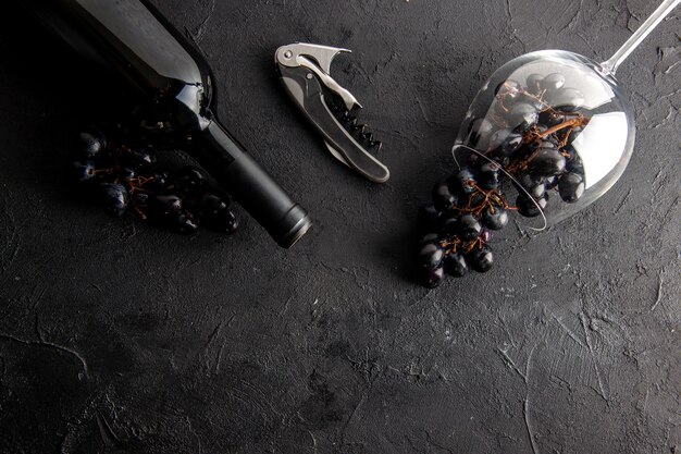 Draufsicht charmante Trauben in umgestürztem Weinglas und Weinflaschen-Weinöffner auf schwarzem Tisch