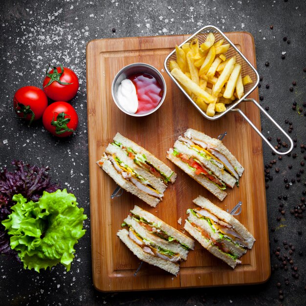 Draufsicht-Club-Sandwich mit Soßenketchup und Mayonnaise und Pommes Frites im hölzernen Servierbrett auf dunklem Steinhintergrund