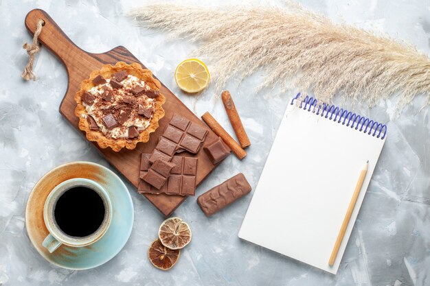 Draufsicht cremiger kleiner Kuchen mit Schokoriegeln Tee-Notizblock und Zimt auf leichtem Schreibtisch süßer Kuchen Zuckercreme-Schokolade