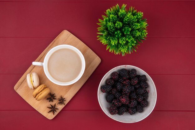 Draufsicht der Brombeere mit einer Tasse Cappuccino mit Macarons auf einem Schneidebrett auf einer roten Oberfläche