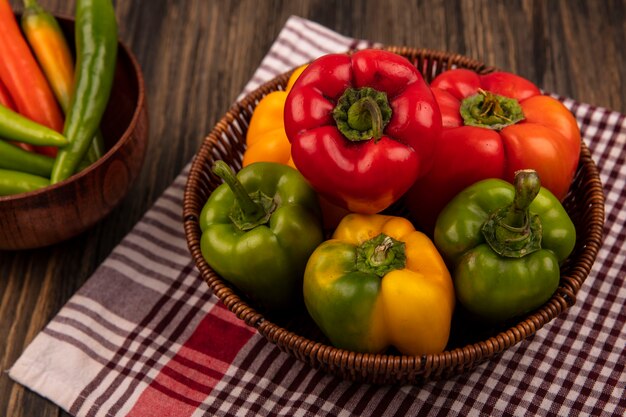 Draufsicht der bunten Paprika auf einem Eimer auf einem karierten Tuch mit lang geformten grünen Paprika auf einer Schüssel auf einer Holzwand