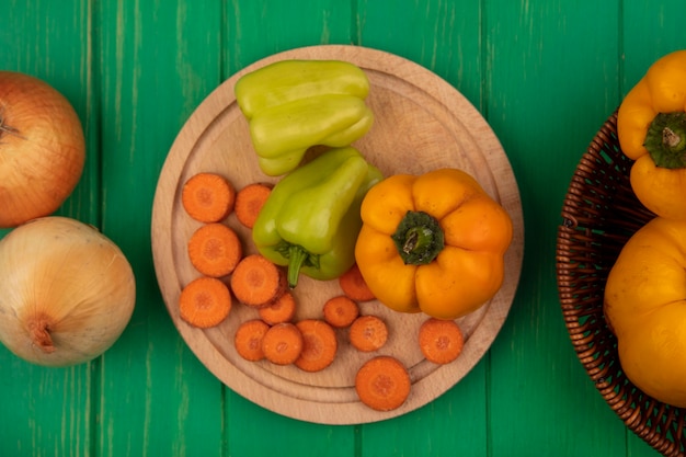 Draufsicht der bunten Paprika auf einem hölzernen Küchenbrett mit gehackten Karotten mit Zwiebeln lokalisiert auf einer grünen Holzwand