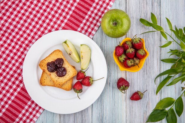 Draufsicht der Erdbeere auf einem weißen Teller mit grünen Äpfeln auf einer rot karierten Tischdecke mit frischen Erdbeeren auf einer gelben Schüssel mit Blättern auf einem grauen hölzernen Hintergrund