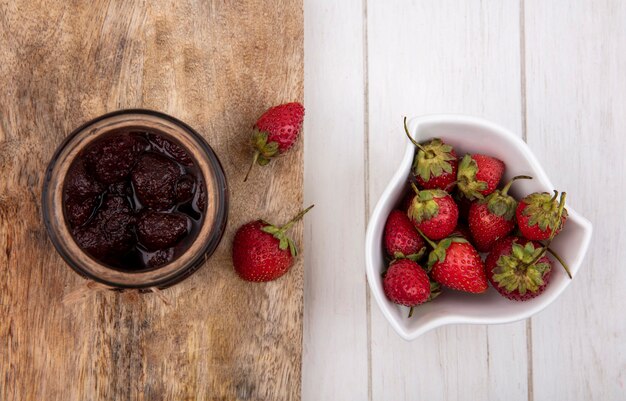 Draufsicht der Erdbeermarmelade auf einem Glas auf einem hölzernen Küchenbrett mit frischen Erdbeeren auf einer weißen Schüssel auf einem weißen hölzernen Hintergrund