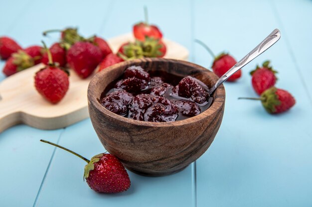 Draufsicht der Erdbeermarmelade auf einer hölzernen Schüssel mit Teelöffel mit frischen Erdbeeren auf einem hölzernen Küchenbrett auf einem blauen Hintergrund