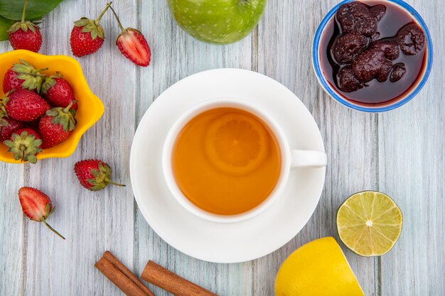 Draufsicht der Erdbeermarmelade auf einer Schüssel mit einer Tasse Tee mit frischen Erdbeeren auf einer gelben Schüssel auf einem grauen hölzernen Hintergrund