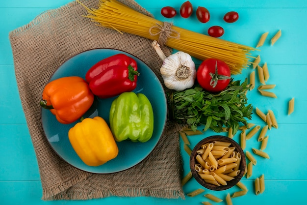 Draufsicht der farbigen Paprika auf einem blauen Teller mit rohen Nudeln und Spaghetti-Kirschtomaten und Knoblauch auf einer türkisfarbenen Oberfläche