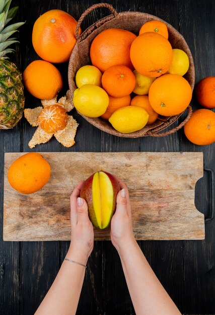 Draufsicht der Frauenhände, die Mango auf Schneidebrett und Zitrusfrüchten als orange Zitronen-Mandarinen-Ananas auf Holztisch halten