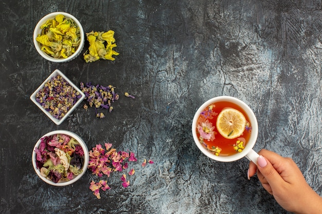 Kostenloses Foto draufsicht der frauenhand, die eine tasse kräutertee und drei schalen der trockenen blumen auf grauem grund hält