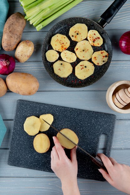 Draufsicht der Frauenhand, die Kartoffel auf Schneidebrett und Pfanne von Kartoffelscheiben Sellerie und Zwiebel auf hölzernem Hintergrund schneidet