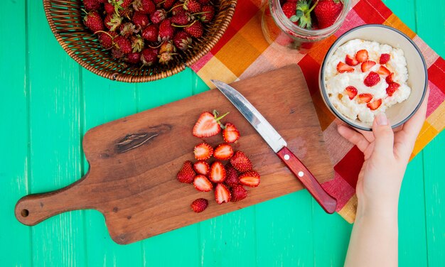 Draufsicht der Frauenhand, die Schüssel Hüttenkäse mit Erdbeeren und Messer auf Schneidebrett und Korb Erdbeeren auf grüner Oberfläche hält