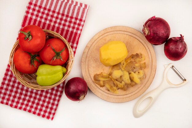 Draufsicht der frisch geschälten Kartoffel auf einem hölzernen Küchenbrett mit Schäler mit Tomaten und Pfeffer auf einem Eimer auf einem karierten Tuch mit roten Zwiebeln lokalisiert auf einer weißen Wand