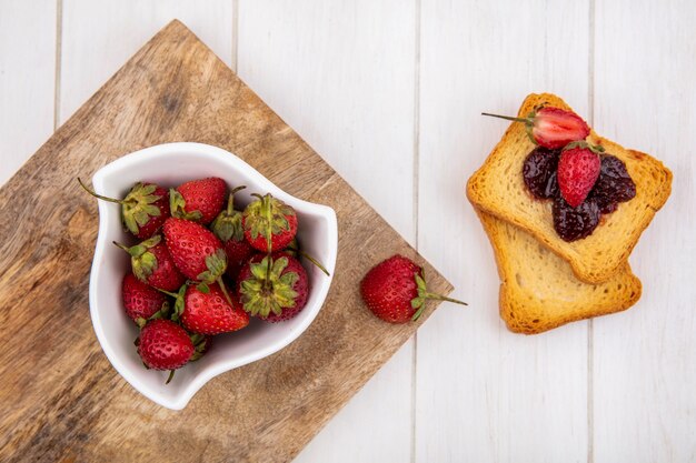 Draufsicht der frischen Erdbeeren auf einer weißen Schüssel auf einem hölzernen Küchenbrett mit gerösteten Brotscheiben auf einem weißen hölzernen Hintergrund