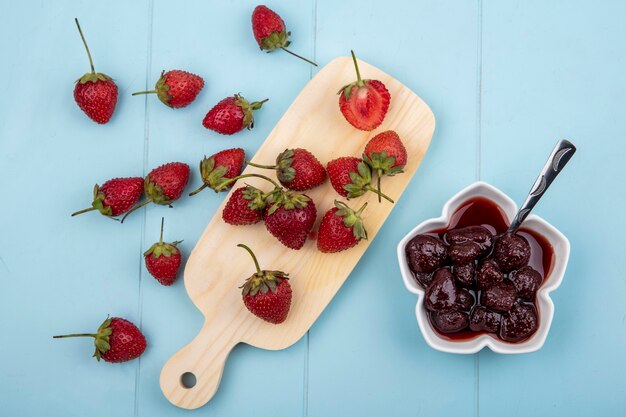 Draufsicht der frischen Erdbeeren lokalisiert auf einem hölzernen Küchenbrett mit einer Erdbeermarmelade auf einer Schüssel auf einem blauen Hintergrund