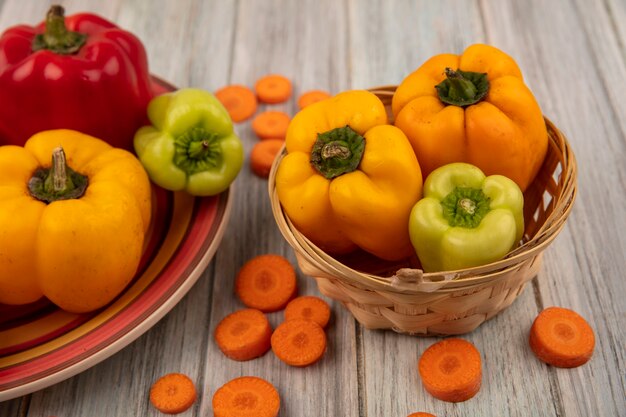 Draufsicht der frischen gelben und grünen Paprika auf einem Eimer mit gehackten Karotten lokalisiert auf einer grauen Holzoberfläche