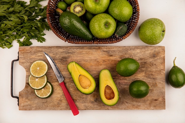 Draufsicht der frischen halben Avocado mit Messer auf einem hölzernen Küchenbrett mit Limetten mit Äpfeln und Kiwi auf einem Eimer auf einem weißen Hintergrund