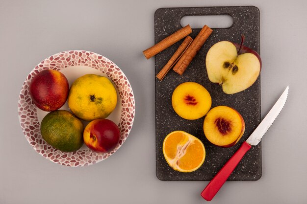 Draufsicht der frischen halben Pfirsiche auf einem schwarzen Küchenbrett mit Mandarine und Apfel mit Messer mit Pfirsichen und Mandarinen auf einer Schüssel auf grauem Hintergrund