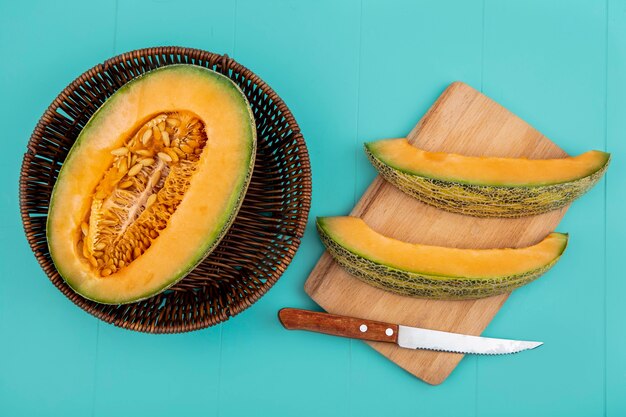 Draufsicht der frischen halbierten Melone Melone auf Eimer mit Melonenscheiben auf Holz Küchenbrett mit Messer auf Blau