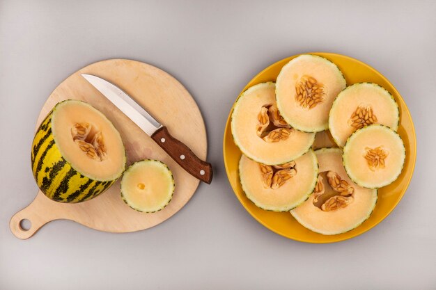 Draufsicht der frischen Melone Melone auf einem hölzernen Küchenbrett mit Messer mit Melonenscheiben auf einem gelben Teller auf einer weißen Wand