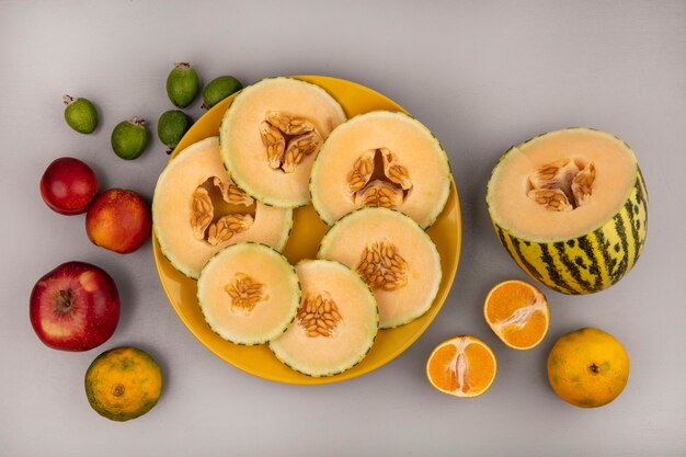 Draufsicht der frischen Melonenscheiben der Melone auf einem gelben Teller mit Apfel-Mandarinen und Feijoas, die auf einer weißen Wand lokalisiert werden