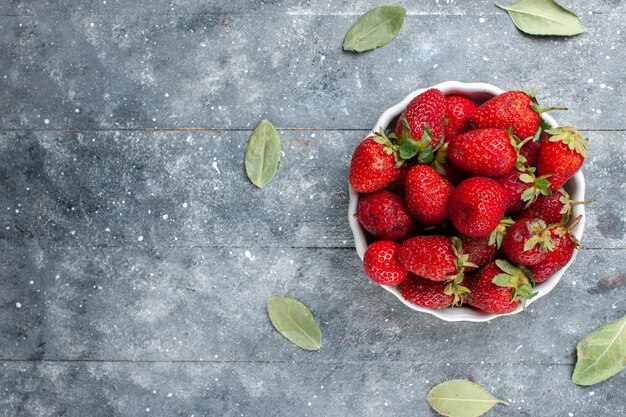 Draufsicht der frischen roten Erdbeeren innerhalb der weißen Platte zusammen mit grünen getrockneten Blättern auf grauer hölzerner, frischer Beerenvitamingesundheit der Frucht