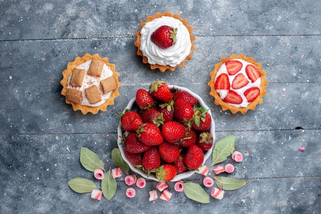 Draufsicht der frischen roten Erdbeeren innerhalb Platte zusammen mit geschnittenen rosa Bonbons und Kuchen auf grauem, frischem Beerenfruchtbeere